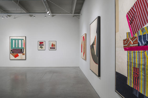 Gallery view displaying framed textile compositions made from hand-cut geometric shapes of silk fabric in jewel tones, patterns, and stripes arranged on white walls. The works feature layered and overlapping fabrics in abstract planar compositions, with areas of translucent and opaque cloth creating depth and movement across the gallery space.