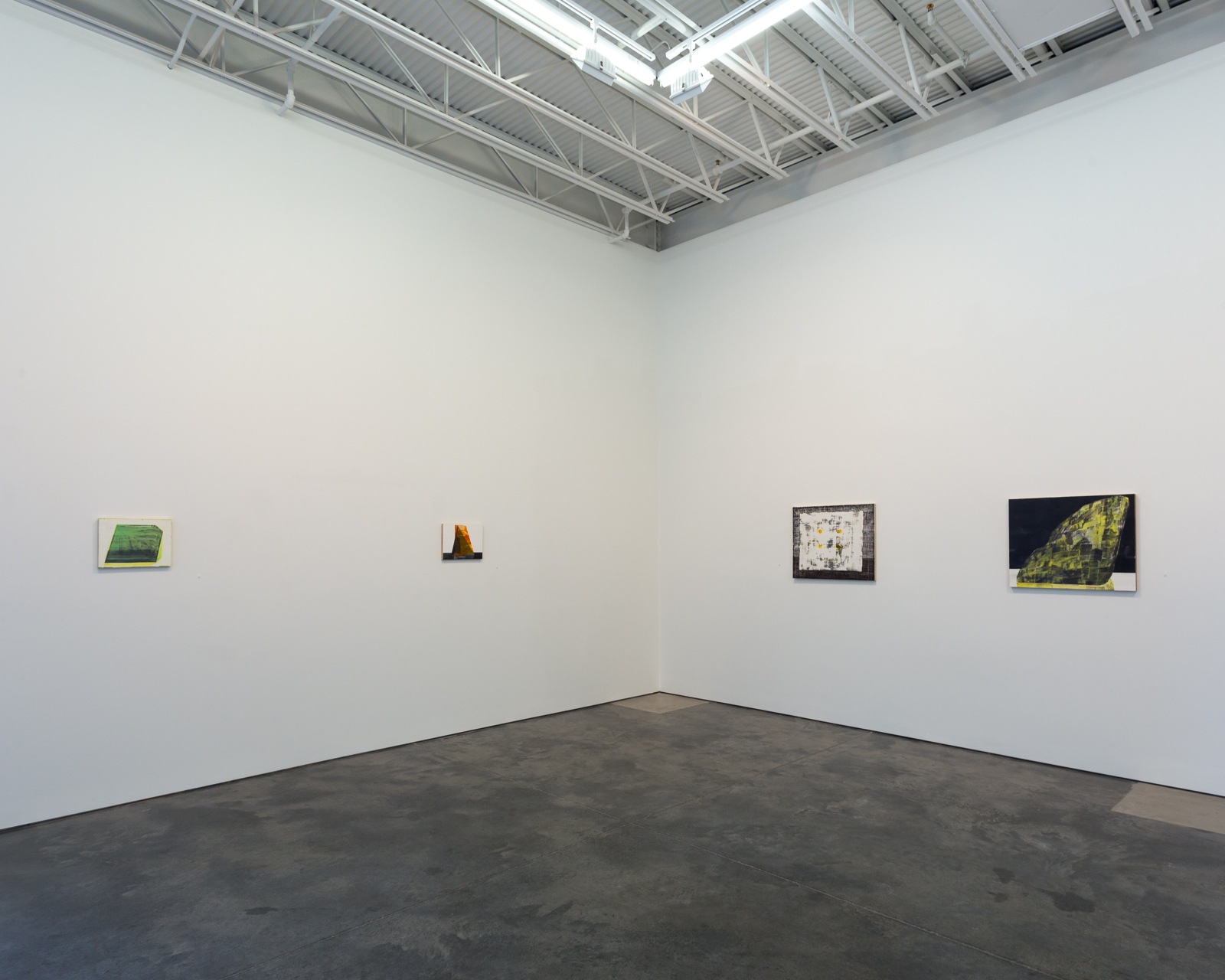 Gallery wall with four small framed works spaced across white walls in a minimalist exhibition space with industrial ceiling. Works feature abstract compositions in green, amber, neutral tones, and dark tones with yellow accents. Polished concrete floor. Spare arrangement emphasizes individual pieces and the architectural void of the gallery.