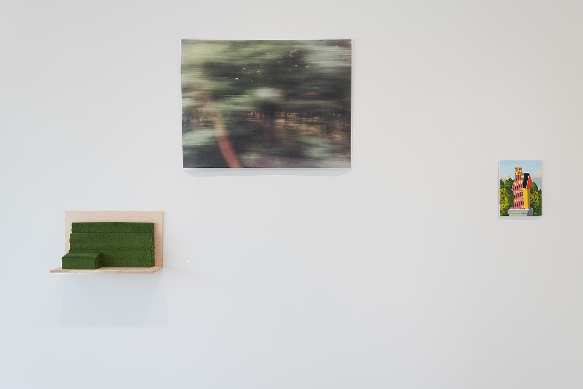 Gallery wall displaying three artworks: a blurred horizontal photograph with green and earth tones mounted centrally, a small wooden shelf with green felt forms on the lower left, and a bright photograph of a colorful striped architectural structure on the right.