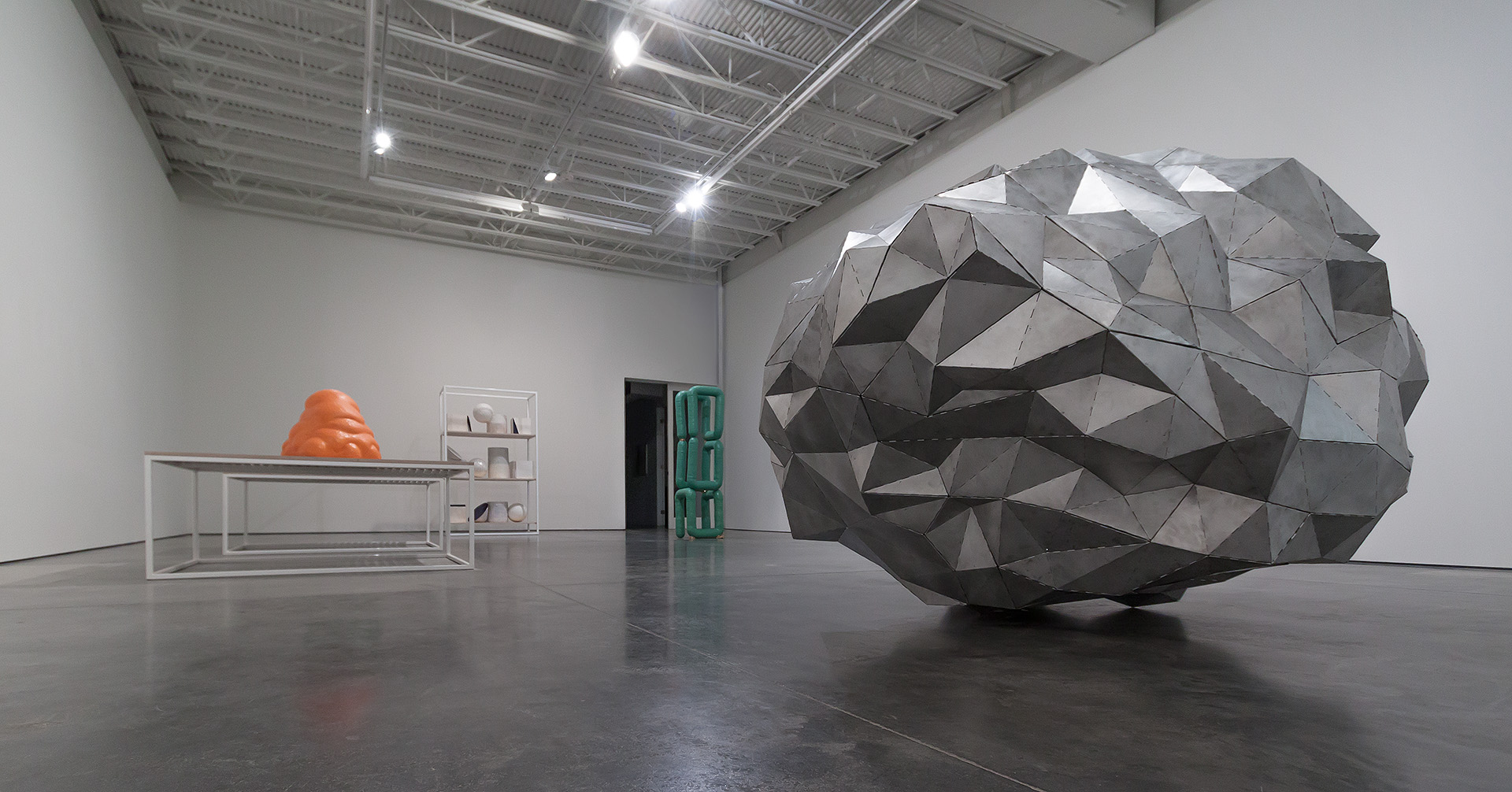 Large-scale geometric sculptural form composed of faceted planes in shades of gray and white dominates the right side of a minimalist gallery space. On the left, a white metal frame table with an orange rounded vessel sits beside a white shelving unit and a green cylindrical object, creating a spare compositional arrangement across the polished concrete floor.