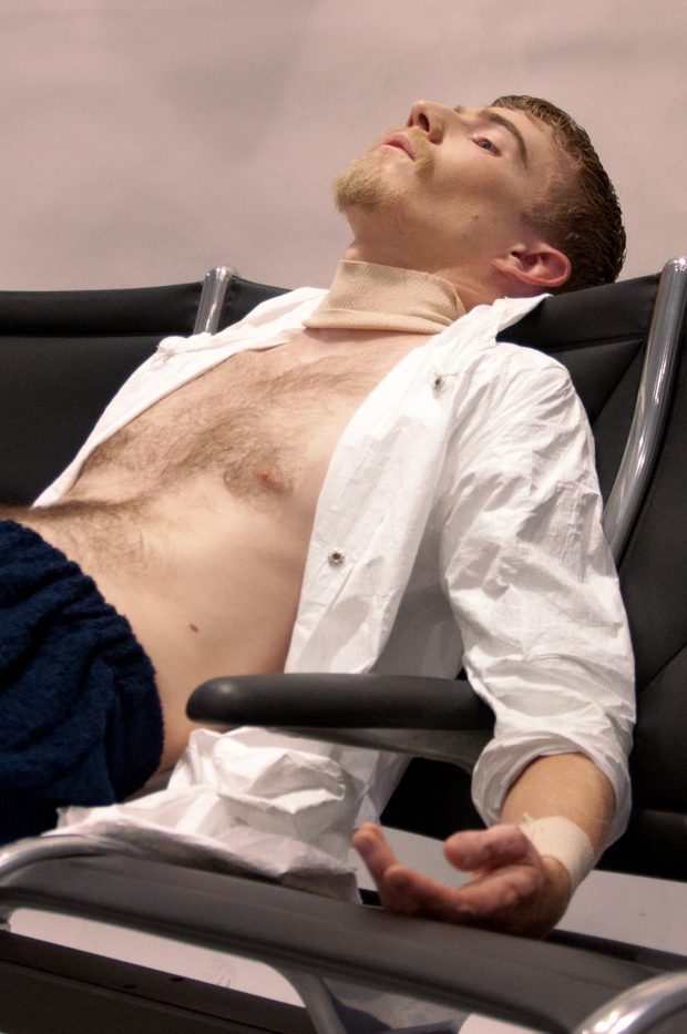 Shirtless man reclined in a black modern chair against a soft pink background, head tilted back with eyes closed in a relaxed pose, wearing an unbuttoned white shirt.