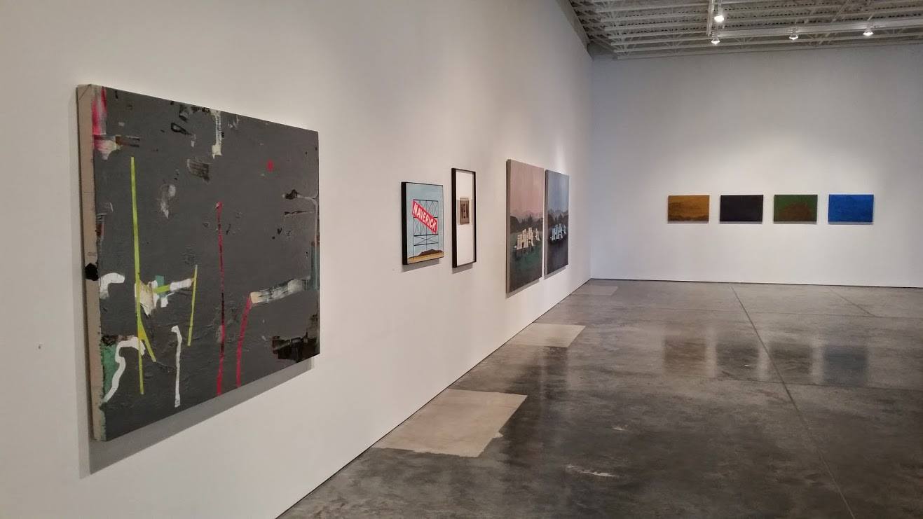 Contemporary art gallery interior displaying multiple works on white walls and concrete flooring. On the left, a large dark canvas with abstract gestural marks in neon yellow, red, and white. Center wall features three framed works including a photograph with red text and two landscape photographs. Right wall shows four small horizontal paintings in warm brown, black, forest green, and cobalt blue tones.