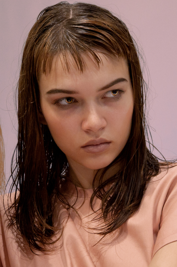 Portrait of a young woman with shoulder-length brown hair and bangs, gazing downward with a contemplative expression against a soft pink background. She wears a pale peach-colored garment and is rendered in photorealistic style with careful attention to facial features and lighting.