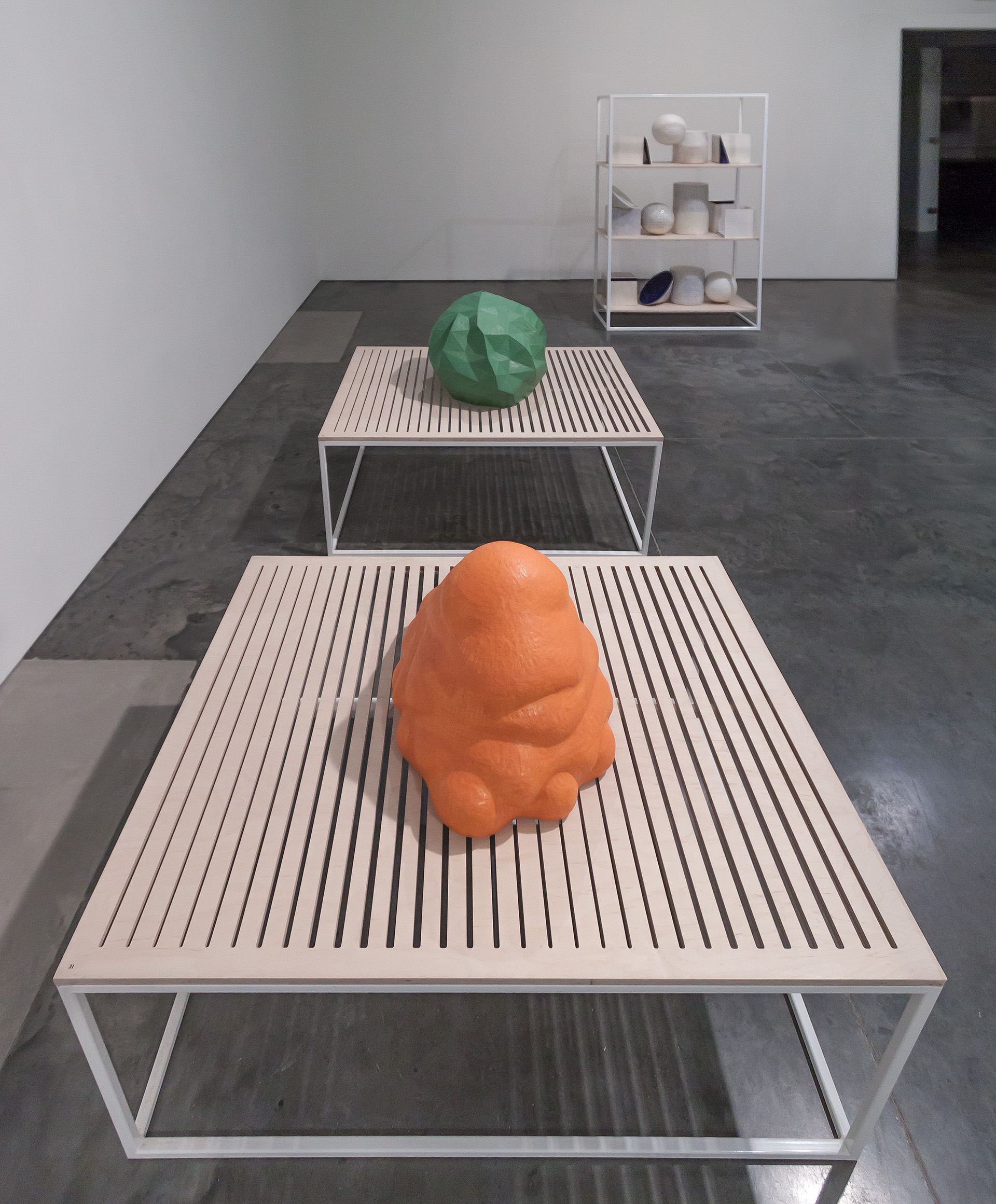 Gallery installation featuring three white metal tables with slatted surfaces arranged in receding perspective on a dark floor. An organic orange sculptural form sits on the nearest table, a geometric green polyhedron on the middle table, and a white shelving unit displaying various ceramic vessels stands in the background.