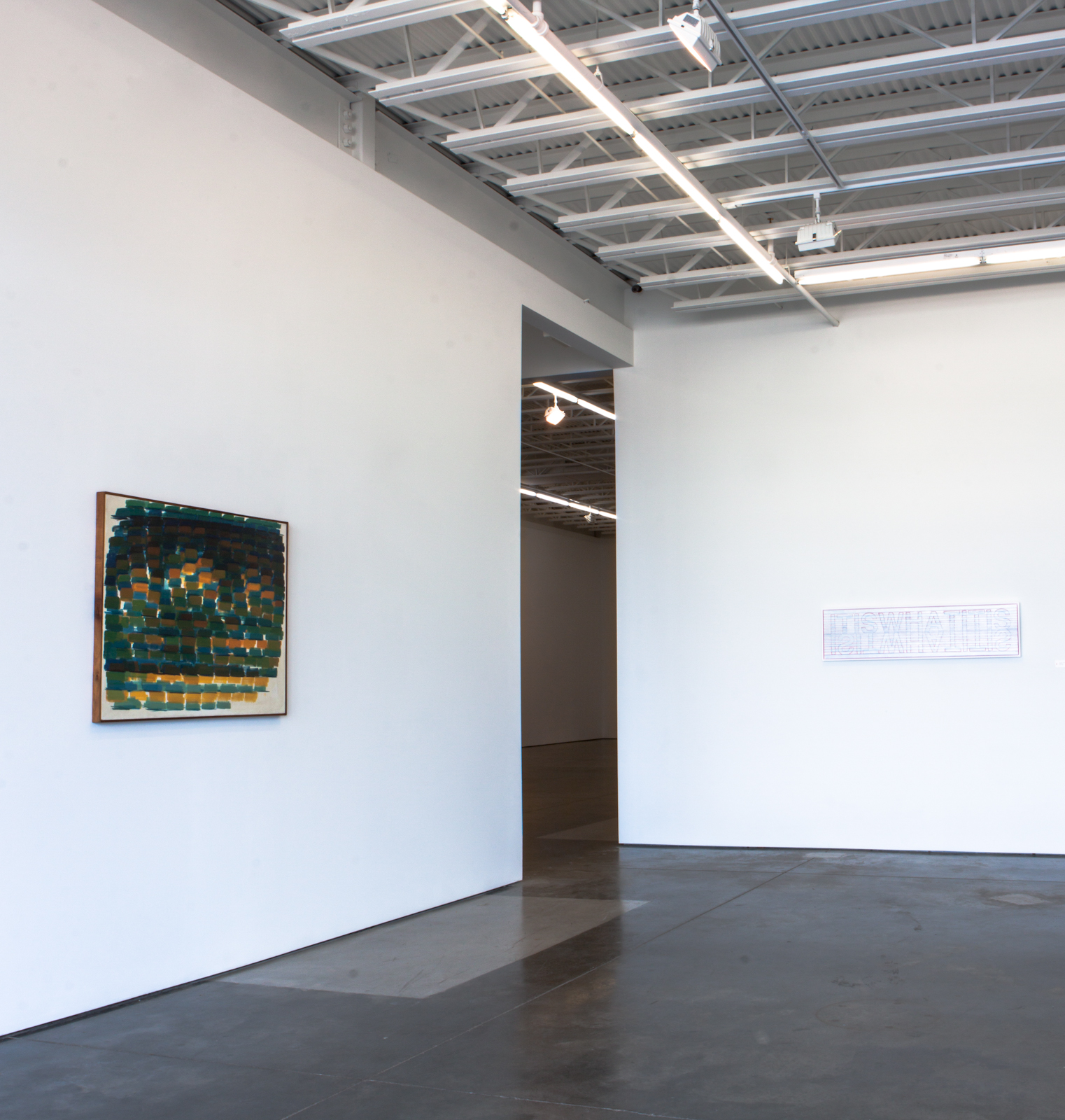 Abstract composition with teal, orange, and yellow geometric shapes arranged in horizontal bands within a wooden frame, displayed on a white gallery wall alongside a small text label and an open doorway leading to an adjacent exhibition space.