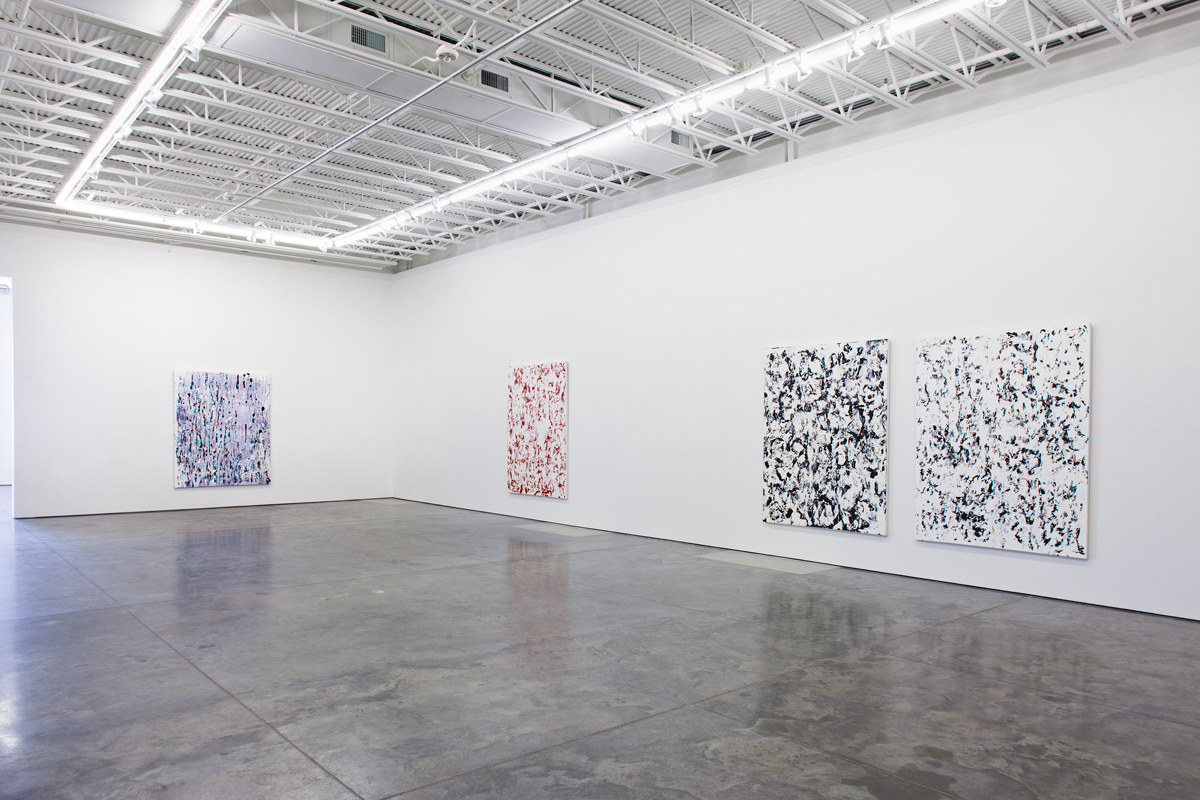 Four large abstract paintings hang on white gallery walls in a minimalist industrial space. The canvases feature gestural, dripped and layered brushwork in varying color combinations—blue and white, red and white, and black and white—creating dense, textured surfaces that fill the entire picture planes.