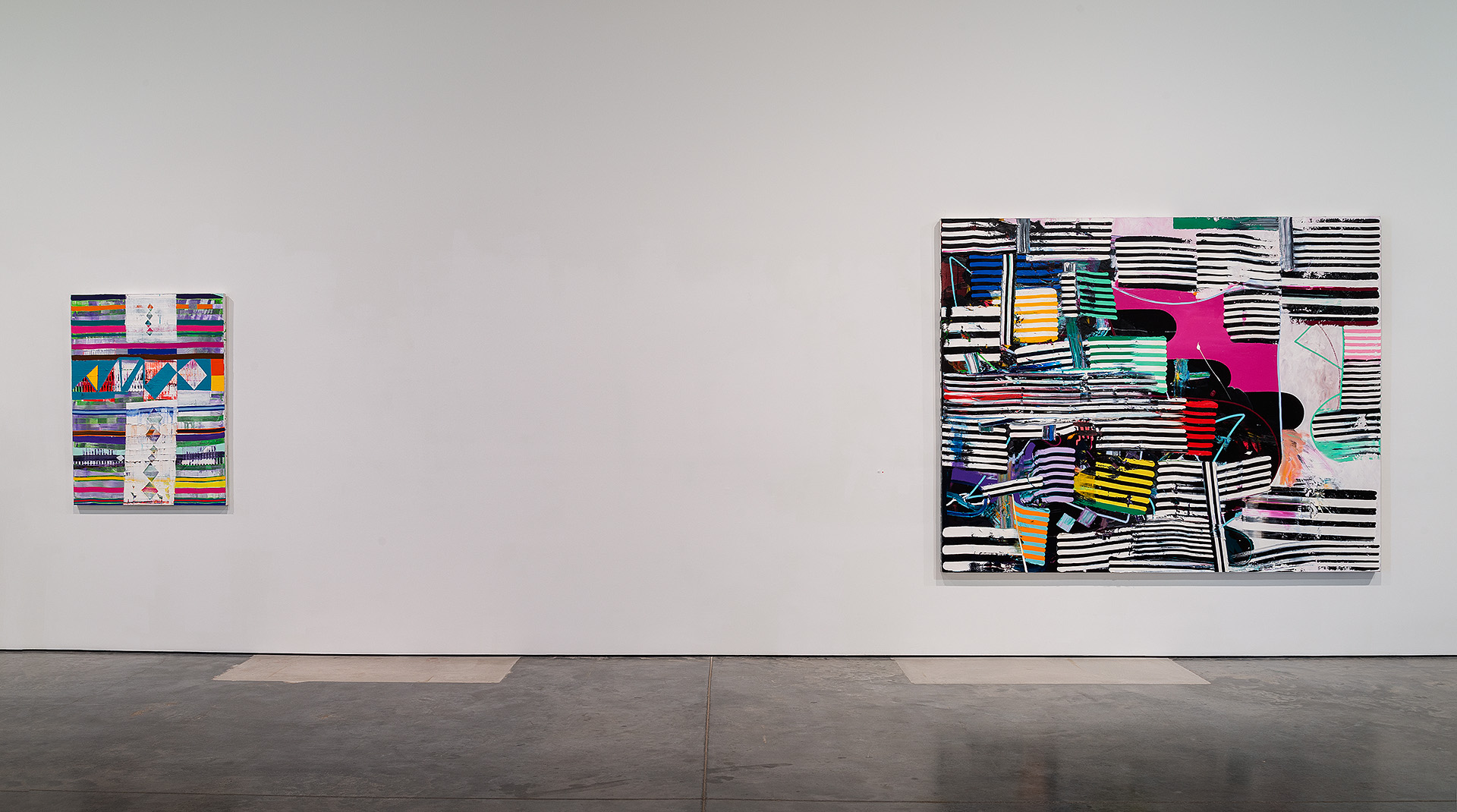 Two abstract paintings hang on a white gallery wall: a smaller multicolored composition on the left featuring horizontal stripes and geometric patterns, and a larger dynamic work on the right with bold black and white striped backgrounds layered with vibrant pink, yellow, blue, and green abstract forms and gestural marks.