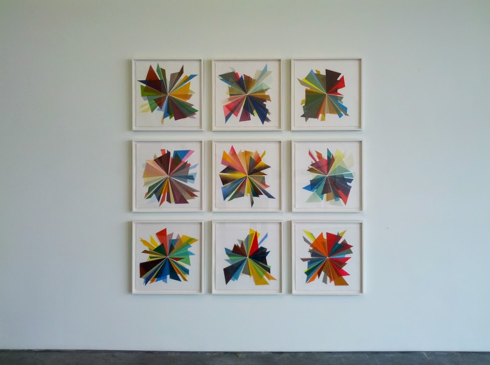 A grid of nine framed works on a white wall, each featuring a radial composition of overlapping triangular and geometric shapes in vibrant colors including red, blue, yellow, green, and burgundy, arranged in starburst patterns that radiate from central points.