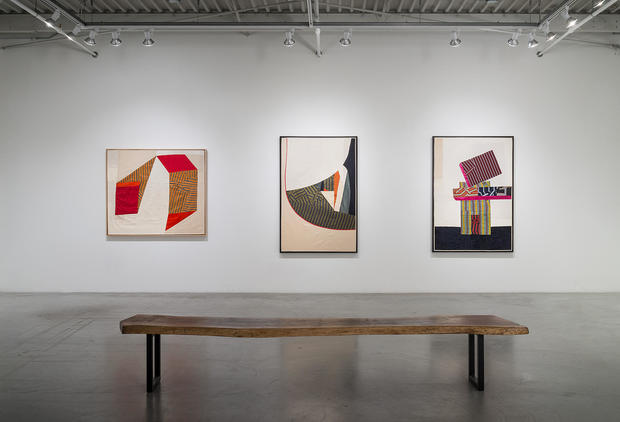 Three framed textile compositions featuring geometric abstract designs created from hand-cut and layered silk fabrics in warm and cool color palettes, hung on a white gallery wall above a wooden bench. The works combine opaque and translucent materials in planar arrangements of overlapping shapes and patterns.
