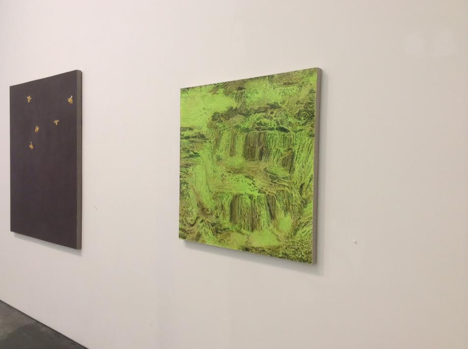 Two paintings hang on a white gallery wall: on the left, a dark charcoal panel with scattered small gold marks; on the right, a bright lime-green canvas depicting an intricate, layered forest or landscape rendered in varying shades of green with dense organic forms and textures.