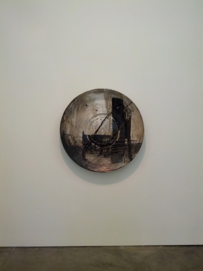 A large circular ceramic vessel mounted on a white gallery wall, its surface marked with gestural black lines and brushstrokes against a weathered, muted gray-brown glaze that suggests depth and movement within the form.
