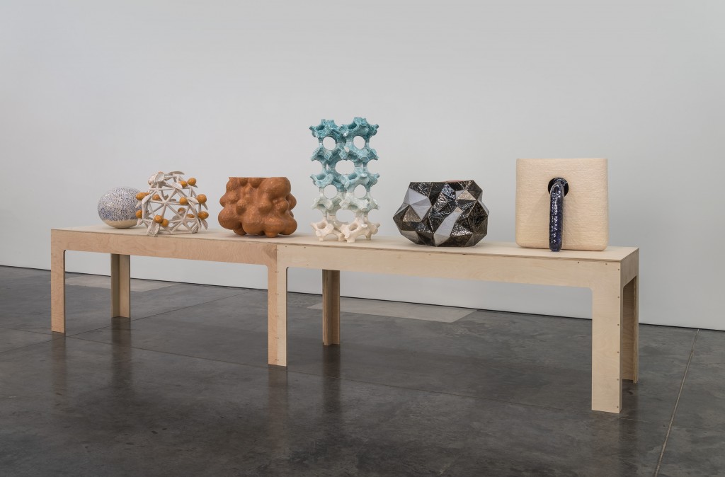 A light wood bench displays five sculptural objects across its surface: a textured spherical form with geometric patterns, an organic brown mass, a stacked turquoise and white geometric structure, a black and white angular form, and a cream-colored rectangular object with a dark cylindrical element. The arrangement is photographed against a white wall and dark floor in a gallery setting.