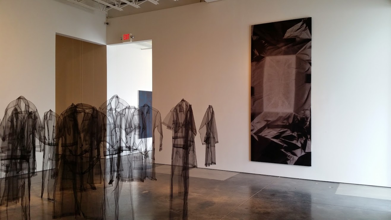 Translucent dark fabric forms stand in a gallery space, appearing as ghostly human silhouettes. A large photograph hangs on the far wall depicting crumpled dark textiles. The installation uses semi-transparent materials to create layered, ethereal figures that interact with light and shadow.