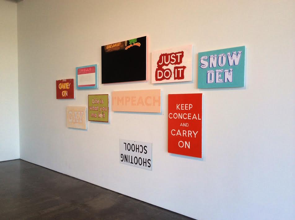 A gallery wall displaying multiple painted signs and panels of varying sizes and colors, including red, turquoise, peach, and white, with text-based messages such as "JUST DO IT," "SNOW DEN," "KEEP CONCEAL AND CARRY ON," and other phrases arranged in an asymmetrical composition against a white wall.