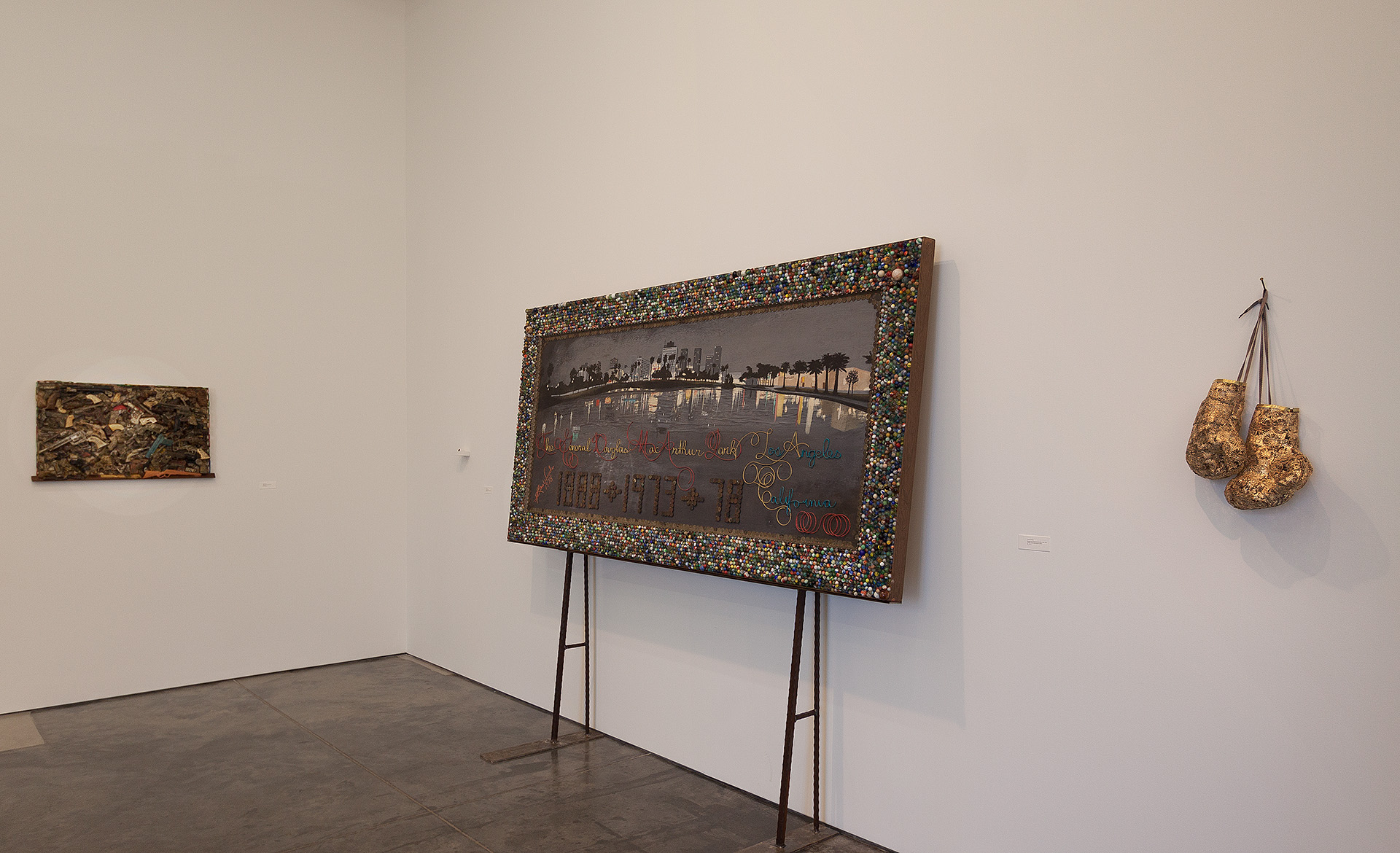 Gallery wall displaying three artworks: a small textured gold-toned piece on the left, a large rectangular work with an ornate beaded frame featuring a cityscape and handwritten text mounted on metal legs in the center, and a pair of gilded sculptural forms suspended on the right wall.