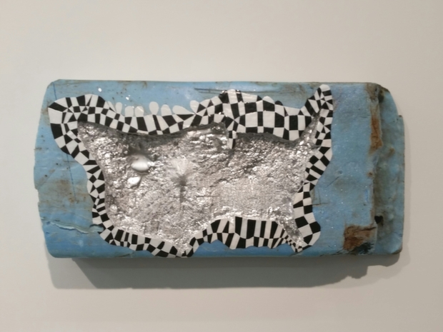 Blue rectangular plaster or concrete panel with a textured gray center surrounded by black and white checkered border patterns, mounted horizontally on a neutral wall.