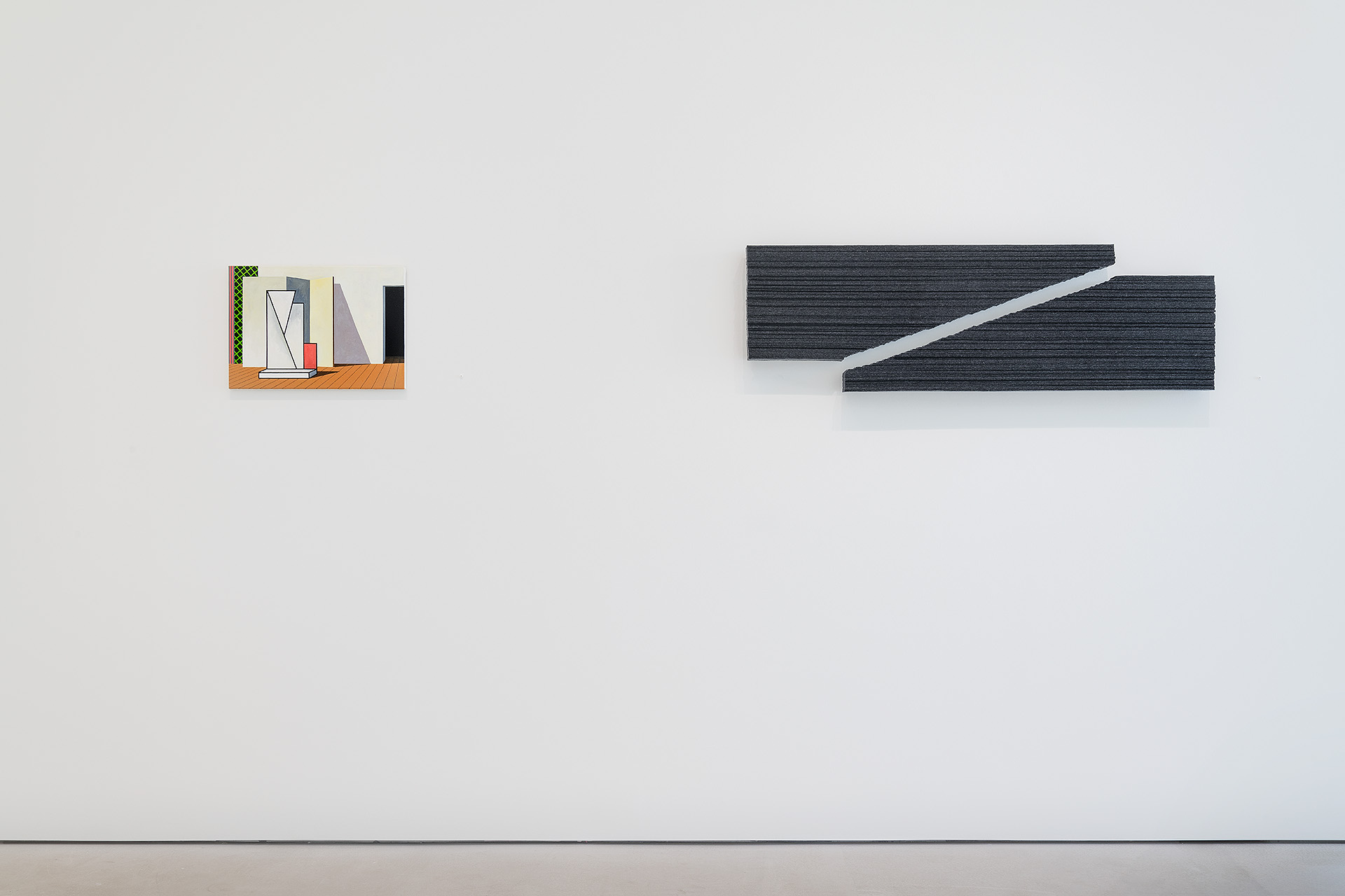 On a white wall, a small illustrated panel depicting interior architectural elements with warm wood tones and geometric forms hangs on the left, while a larger dark gray sculptural object with angular cut-outs and a white diagonal element extends from the right side.