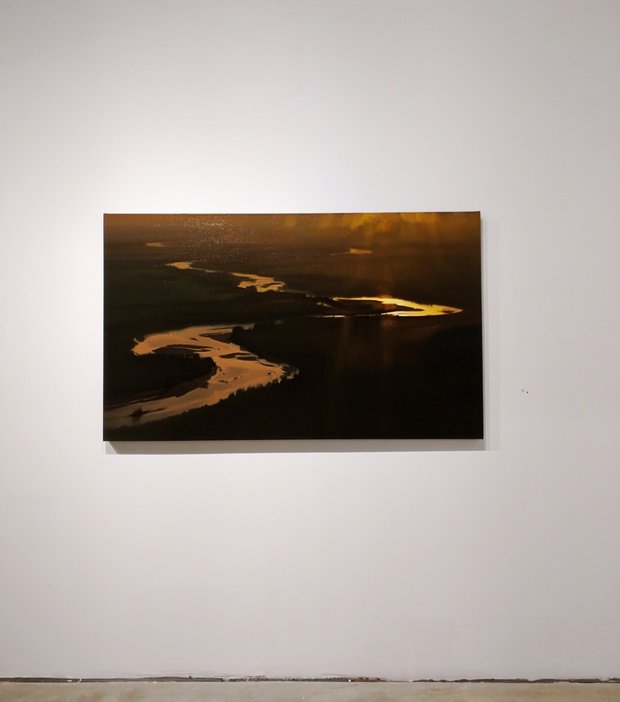 Aerial photograph of a winding river at dusk, its meandering curves illuminated in golden light against dark surrounding landscape, mounted on a white gallery wall.