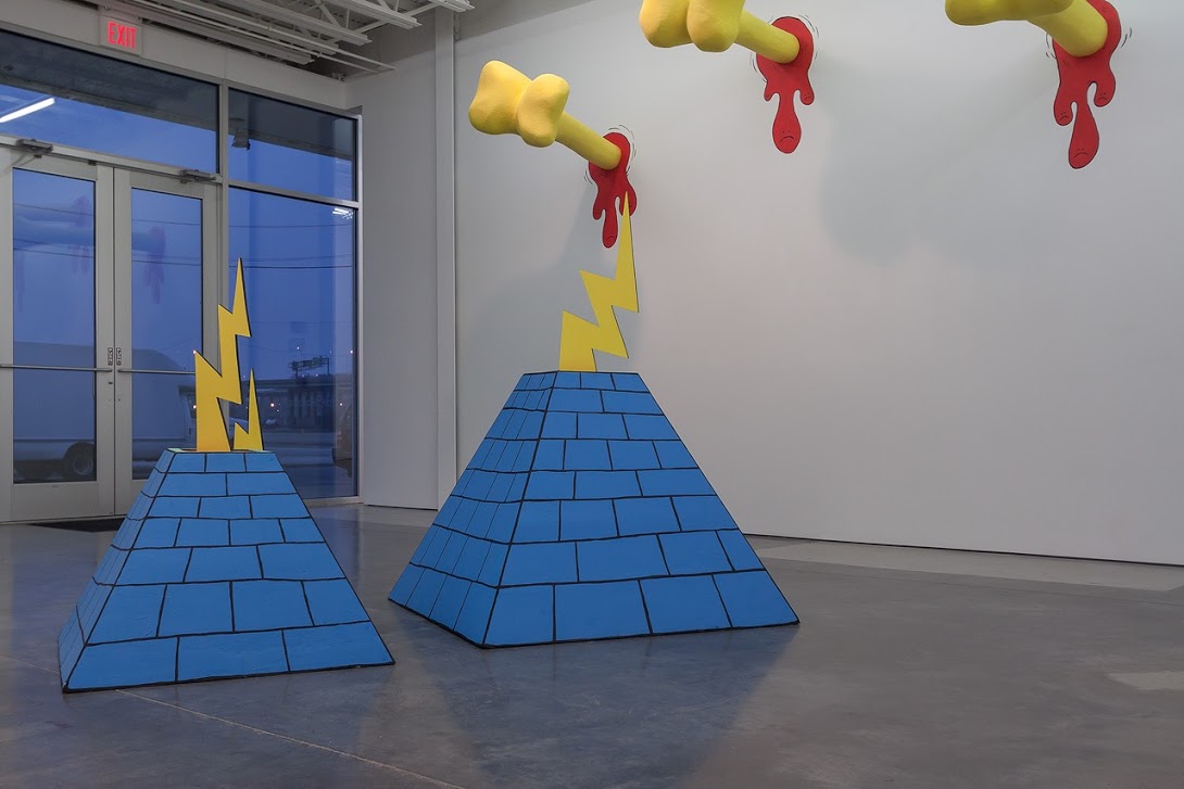 Two large blue pyramidal structures with grid patterns sit on a polished concrete floor beneath hanging yellow and red sculptural forms dripping downward. Lightning bolt shapes in yellow emerge from the pyramids' tops, creating a dynamic, cartoonish composition in a minimalist gallery space with floor-to-ceiling windows.