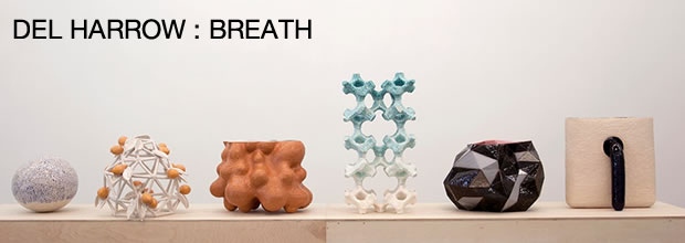 Six sculptural forms arranged in a line on a neutral shelf: a textured white spherical form, an orange geometric lattice structure, a brown organic bulbous shape, a pale blue-green crystalline tower, a dark abstract curved form, and a cream-colored cubic piece with a black handle. The works demonstrate varied materials and surface qualities, from smooth to intricate, exploring themes of volume and repetitive form.