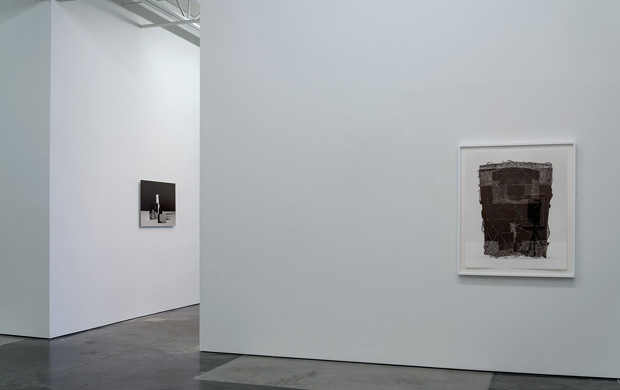 Gallery interior with two framed artworks on white walls: a small dark abstract photograph on the left wall, and a larger textured brown drawing or print on the right wall, displayed against a polished concrete floor.
