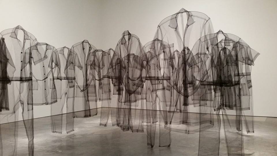 Translucent mesh forms resembling business suits and coats overlap and layer across a gallery floor, creating ghostly silhouettes that fade in and out of focus. The monochromatic figures stand in a circular arrangement, their semi-transparent fabric suggesting absence and invisibility.
