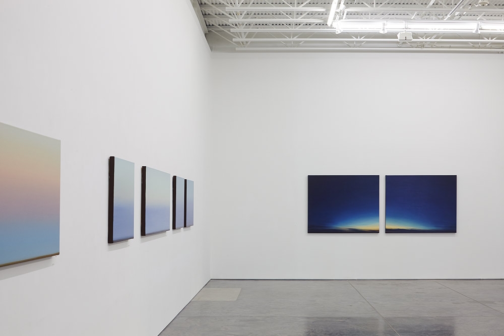 Gallery installation view showing five framed landscape paintings arranged on white walls. On the left wall hangs a single large canvas with soft pink and blue gradient tones. The center wall displays four vertical canvases in a row, each depicting subtle atmospheric gradations from light to dark blue. The right wall features two horizontal canvases side by side with deep blue skies transitioning to warm golden and orange tones along the horizon. The paintings emphasize luminous color shifts and atmospheric perspective characteristic of twilight and dawn phenomena.