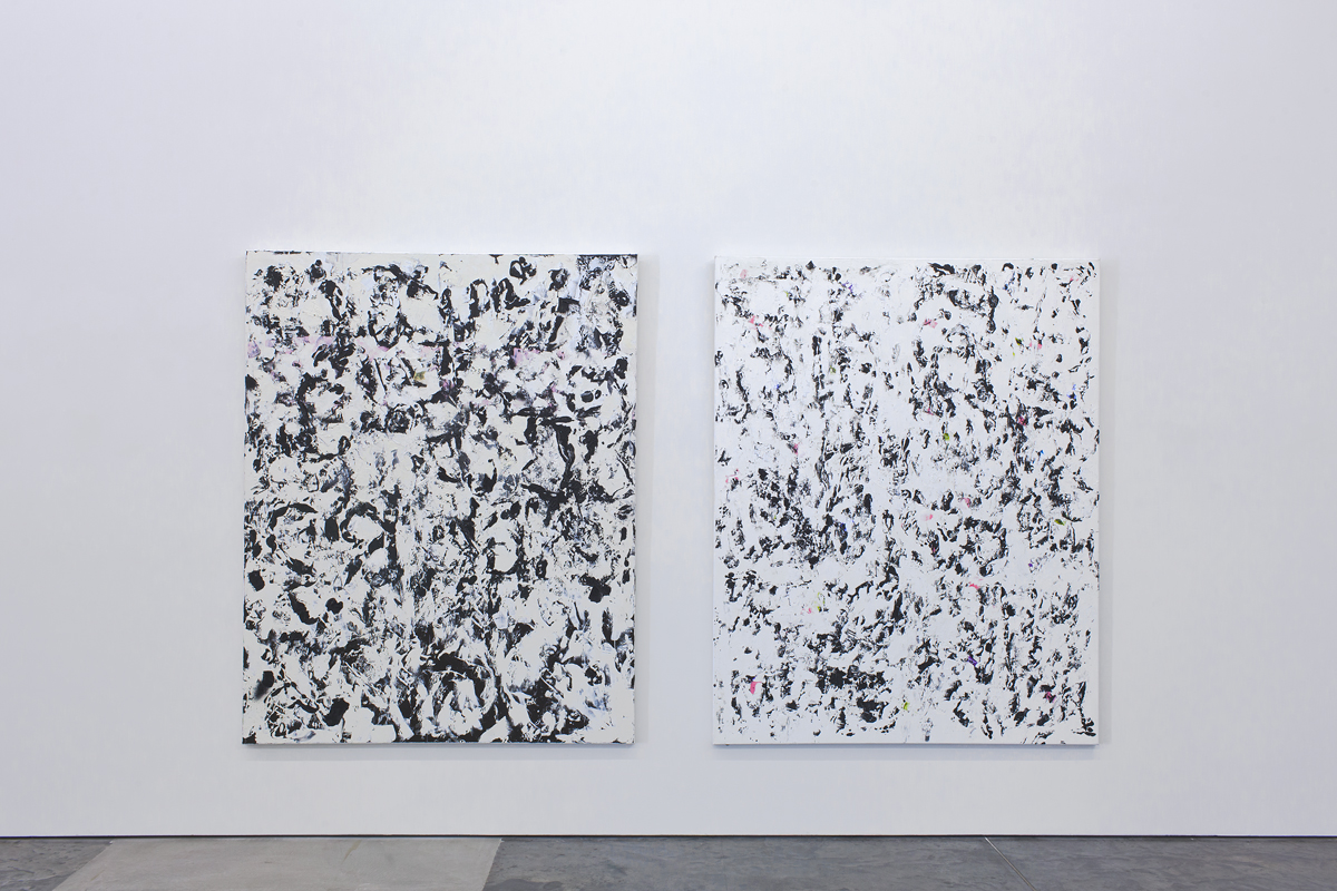 Two large abstract paintings with white backgrounds densely covered in black gestural marks, drips, and fragmented brushstrokes that create an all-over composition reminiscent of action painting. The works hang side by side on a gallery wall, their layered, energetic surfaces suggesting emotional intensity through accumulated marks and splatters.