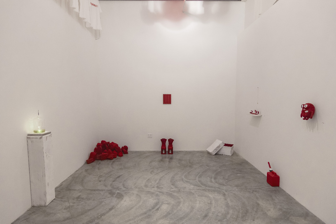 A sparse gallery installation with a gray concrete floor features various red objects and materials scattered throughout a white-walled room: a pile of red fabric, cylindrical red forms, a red shovel, wall-mounted red sculptures, and a white pedestal with a glass vessel on the left side.