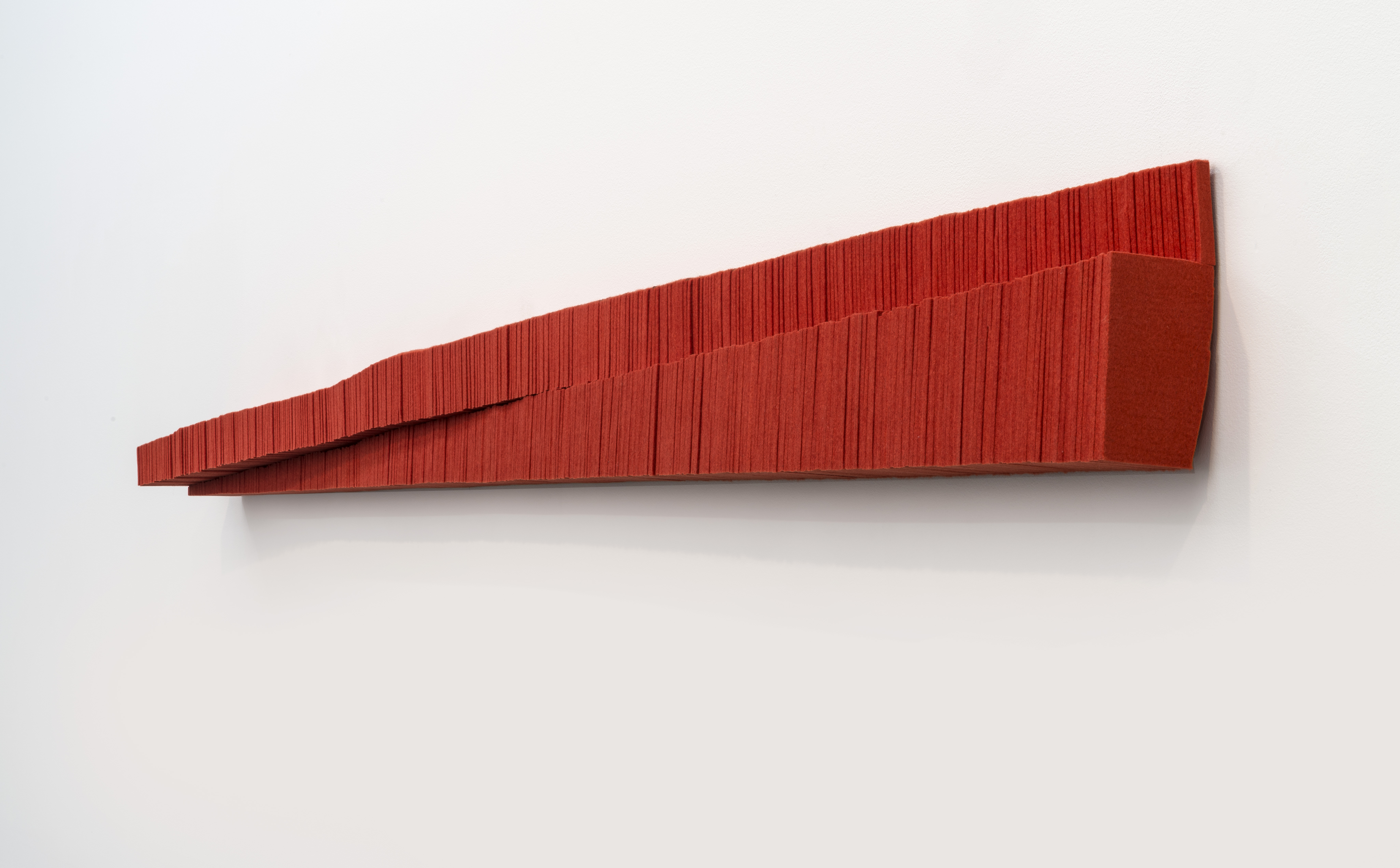 Red industrial felt sculpture mounted horizontally on a white wall, featuring a tapered wedge form with deeply textured vertical striations across its surface, creating a linear pattern that emphasizes mass and shadow.