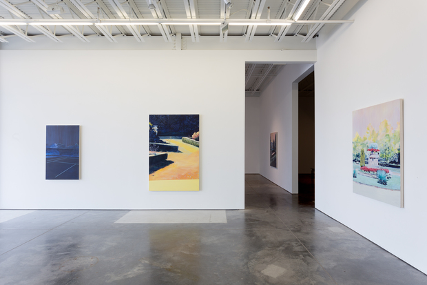A contemporary art gallery interior with polished concrete floors and white walls displays four colorful paintings at varying heights. Works feature vibrant compositions in blues, yellows, greens, and pinks, showcasing constructed landscapes and architectural spaces rendered in bold, contrasting colors with crisp geometric forms.