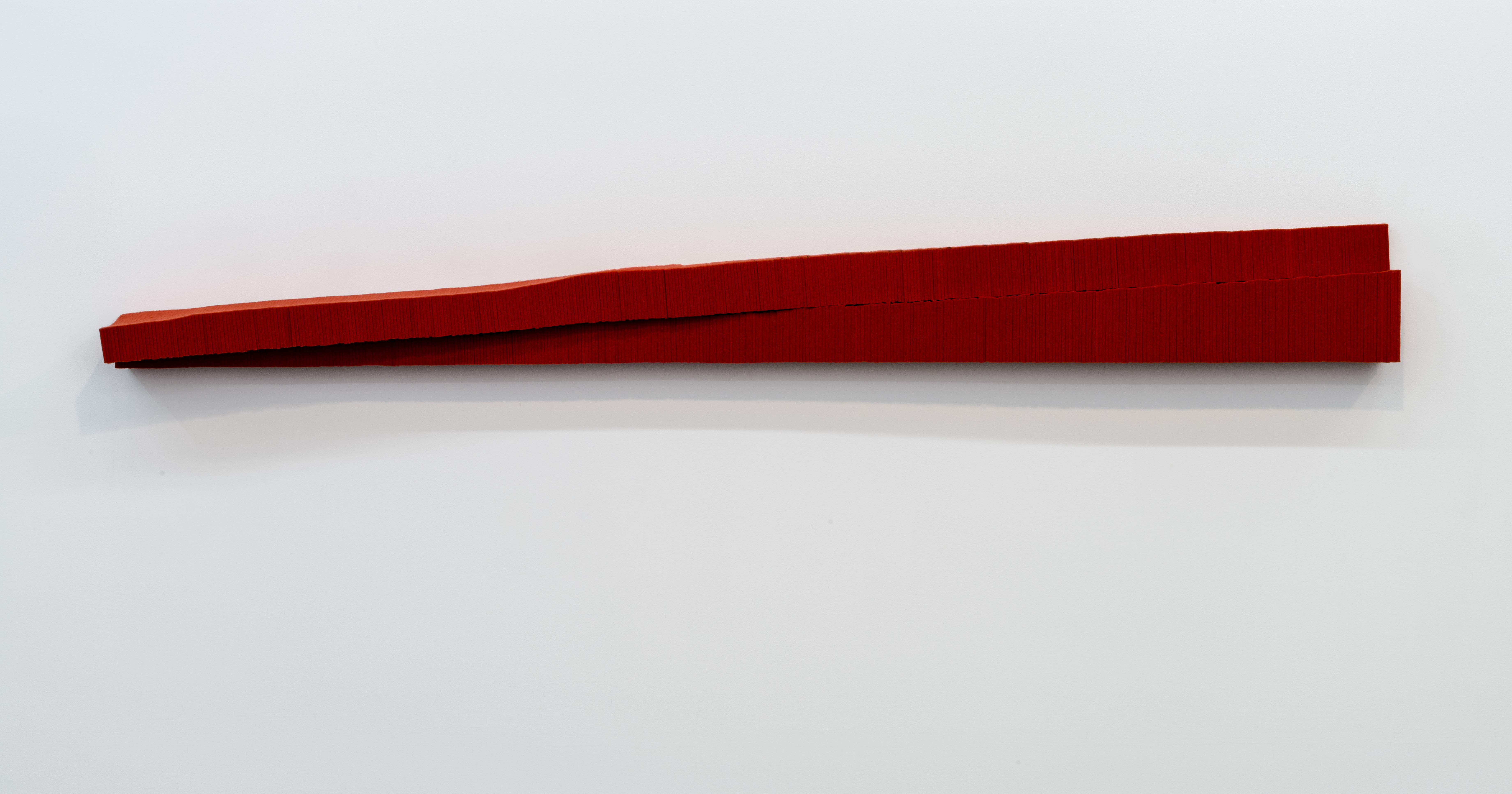 A deep red rectangular form extends horizontally across a light gray wall, its surface appearing densely compressed with subtle textural variations suggesting industrial felt or similar material. The work casts a defined shadow, emphasizing its three-dimensional mass and austere, minimalist presence.