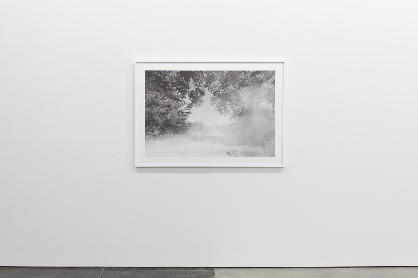Black and white photograph in a white frame depicting a misty rural landscape with dense trees framing the edges and fog obscuring a gravel road or clearing in the center. The image has a soft, atmospheric quality with minimal contrast between sky and land.