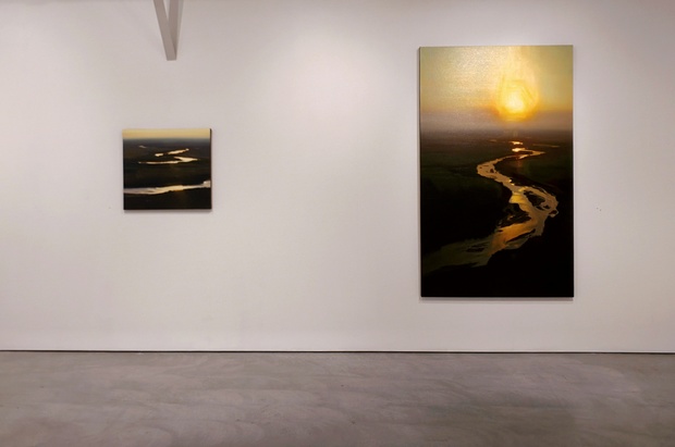 Two photographs mounted on a white gallery wall depicting aerial views of a meandering river at sunrise or sunset. The smaller work on the left shows a horizontal band of golden light reflecting off dark water, while the larger vertical composition on the right captures a winding river's luminous curves cutting through shadowed landscape beneath a glowing sun.