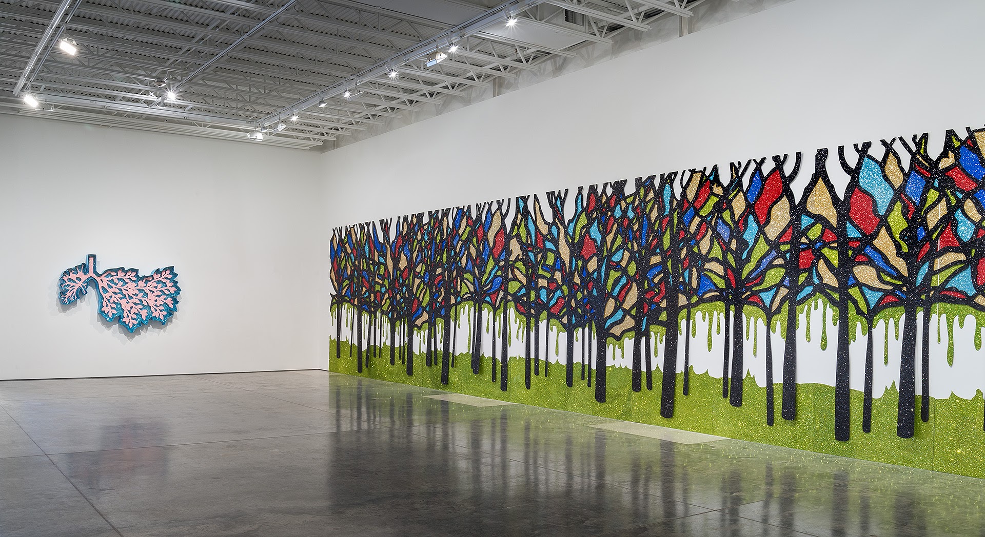 A large-scale wall installation depicting a forest of stylized trees with black trunks, green grass base, and luminous canopies filled with stained-glass-like patterning in vibrant blues, reds, yellows, and greens, adorned with shimmering glitter. To the left hangs a smaller coral-pink and blue branching form suggesting biological or anatomical origins.