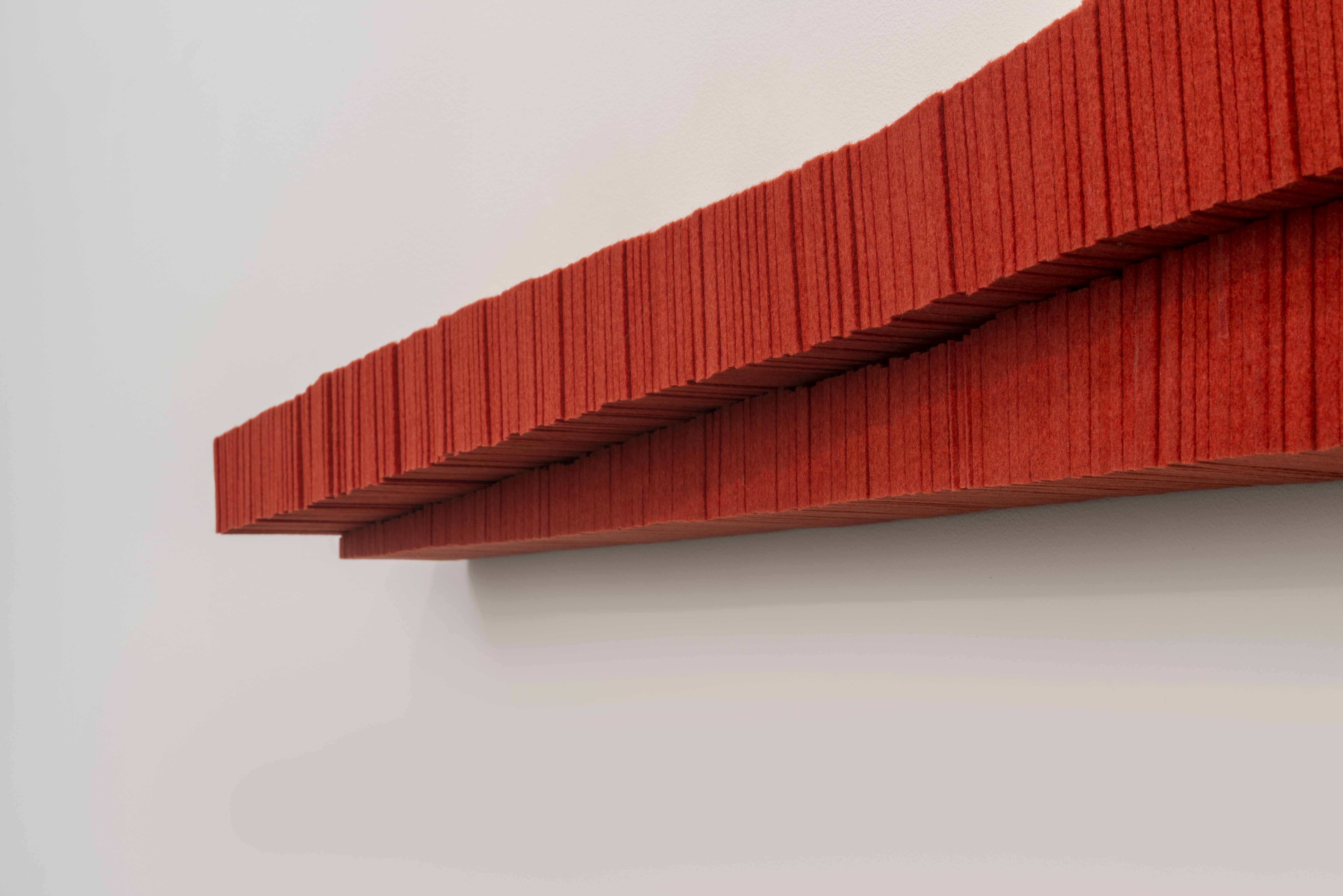 A long, horizontally-oriented red sculptural form with deeply textured vertical ridging extends across a white wall, casting a dramatic shadow. The saturated red material appears dense and industrial, with visible linear striations creating a rhythmic pattern across its surface.