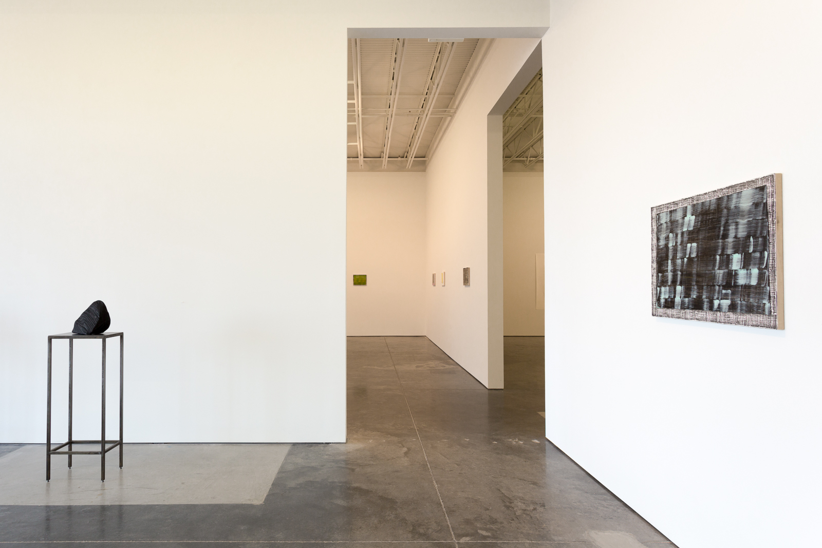 Contemporary gallery interior with polished concrete floors featuring a tall metal pedestal on the left supporting a black sculptural form, a central white architectural partition with recessed elements, and a textured abstract artwork mounted on the right wall with blue and white geometric patterns.