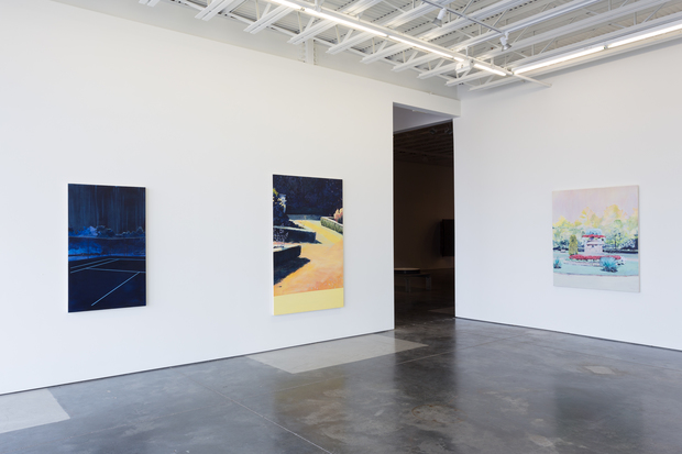 Contemporary gallery interior with three paintings hung on white walls and a dark doorway. Left wall features a dark navy abstract composition with geometric forms; center wall displays a yellow and navy landscape-like scene; right wall shows a pastel-toned garden or pool scene. Polished concrete floor, industrial ceiling with exposed beams and skylights.