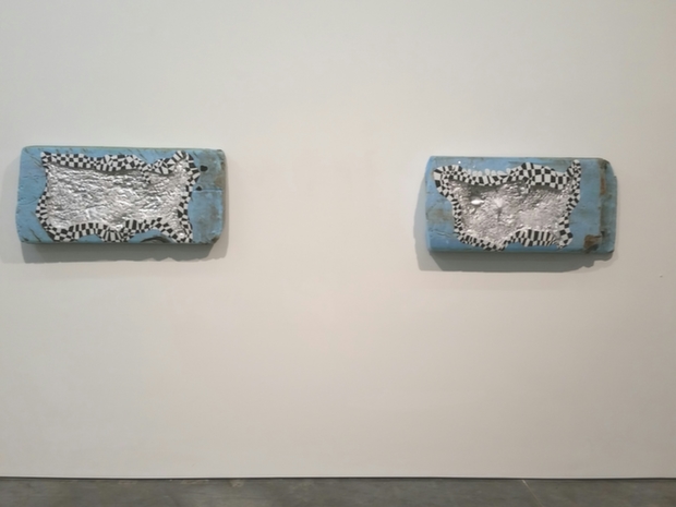 Two rectangular blue-gray wall-mounted panels with silver-textured central relief forms featuring intricate dotted and linear surface patterns, positioned symmetrically on a white gallery wall.