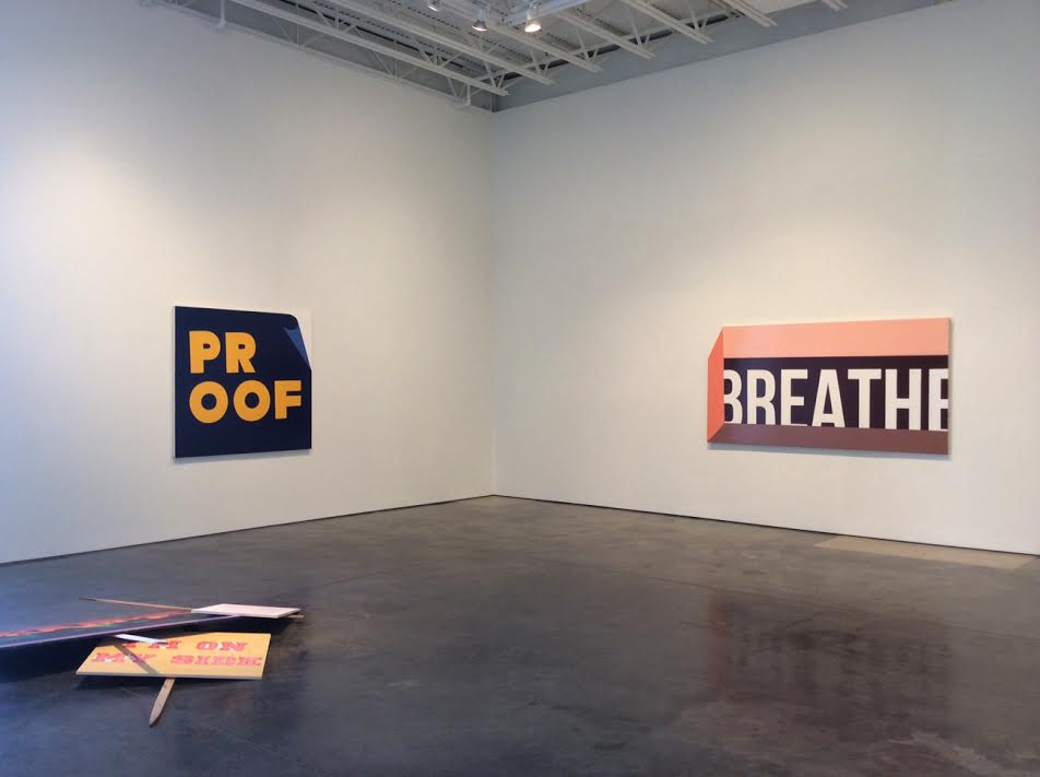 Gallery corner featuring two large wall-mounted signs with bold typography in navy blue and coral orange, each with partially obscured text reading "PROOF" and "BREATHE." A third sculptural element lies on the dark concrete floor in the foreground. Minimalist installation emphasizing layered meanings through fragmented words and strategic cropping.