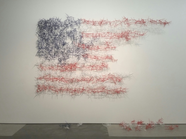 Abstract wall installation composed of densely tangled dark blue wire forming a gestural head or profile on the left side, transitioning into sweeping horizontal bands of red wire that extend across the white wall like fragmented stripes. Loose red wire elements cascade onto the gray floor below, creating a sense of movement and decay.