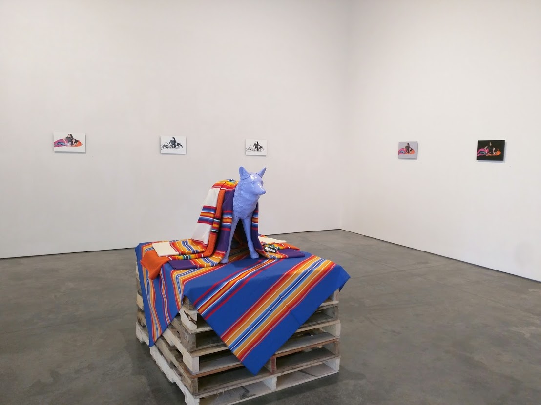 A blue sculptural figure of a horse stands on a wooden pallet draped with vibrant striped blankets in blue, orange, red, and yellow. Five framed photographs hang on the white gallery wall behind the installation. The composition sits on a polished concrete floor.