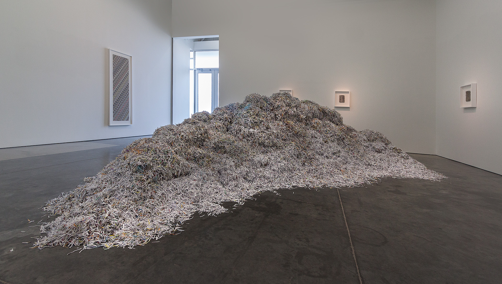 A large mound of shredded paper and cardboard fragments dominates the center of a white-walled gallery space with dark flooring. Small framed works hang on the walls in the background, with natural light streaming through a doorway. The accumulated material appears to be the result of systematic collection and deconstruction, creating an imposing sculptural form through repetition and layering of everyday refuse.