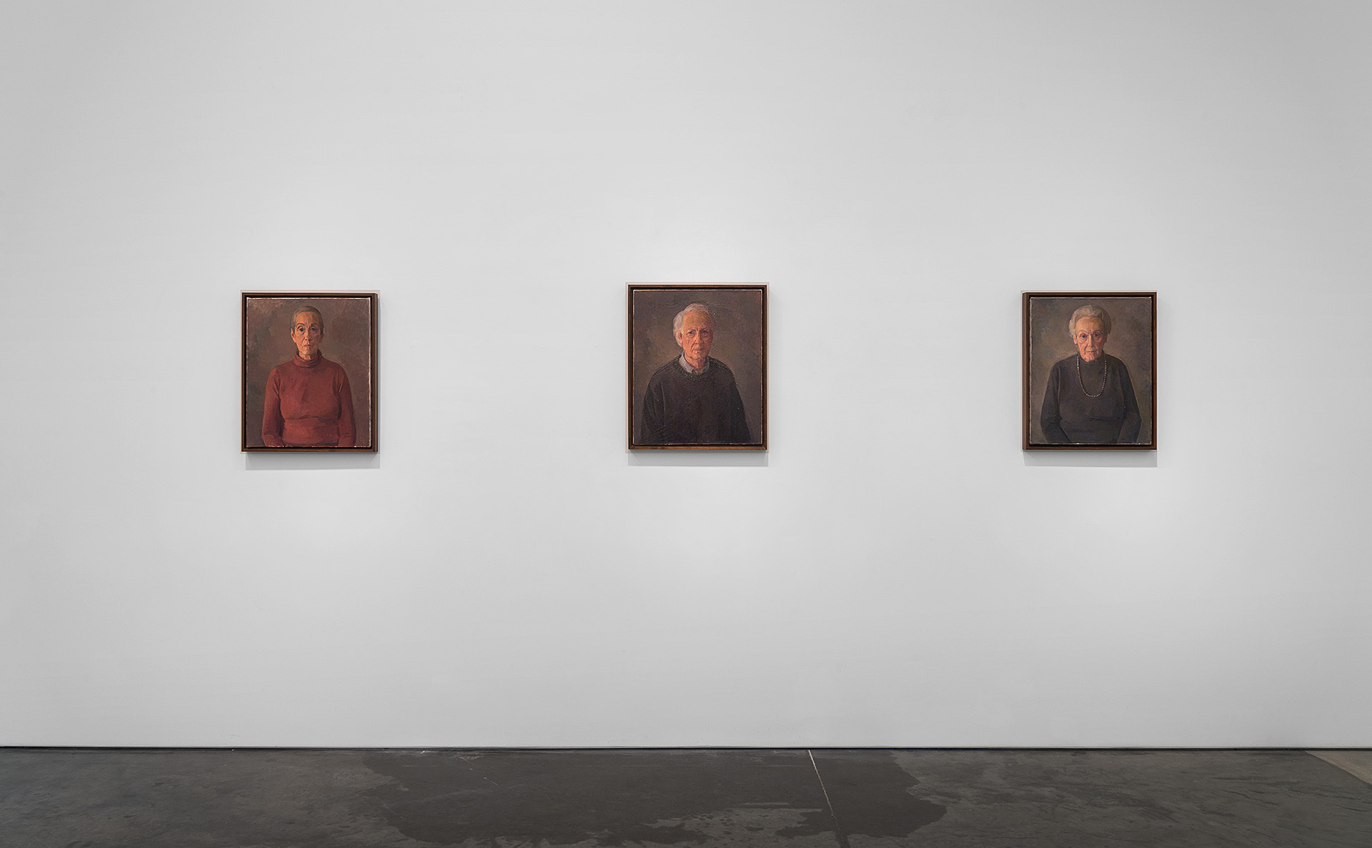 Three framed portrait paintings hung horizontally across a gallery wall, each depicting a solitary figure in muted earth tones against dark backgrounds. The left portrait shows a figure in a red garment, while the center and right figures wear darker clothing. The paintings feature a restrained, contemplative style with warm, subdued lighting that emphasizes the subjects' faces.