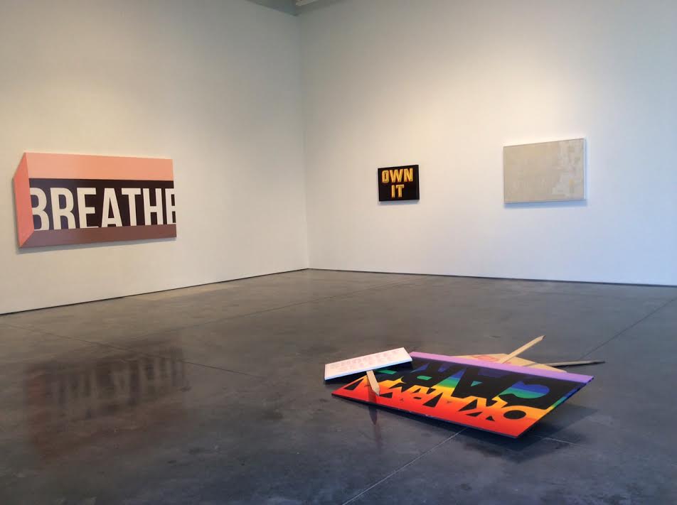 Contemporary gallery installation featuring three wall-mounted text-based artworks in bold typography against white walls, with a colorful foam board construction on the dark floor displaying layered text in rainbow and black sections, accompanied by white paper elements.