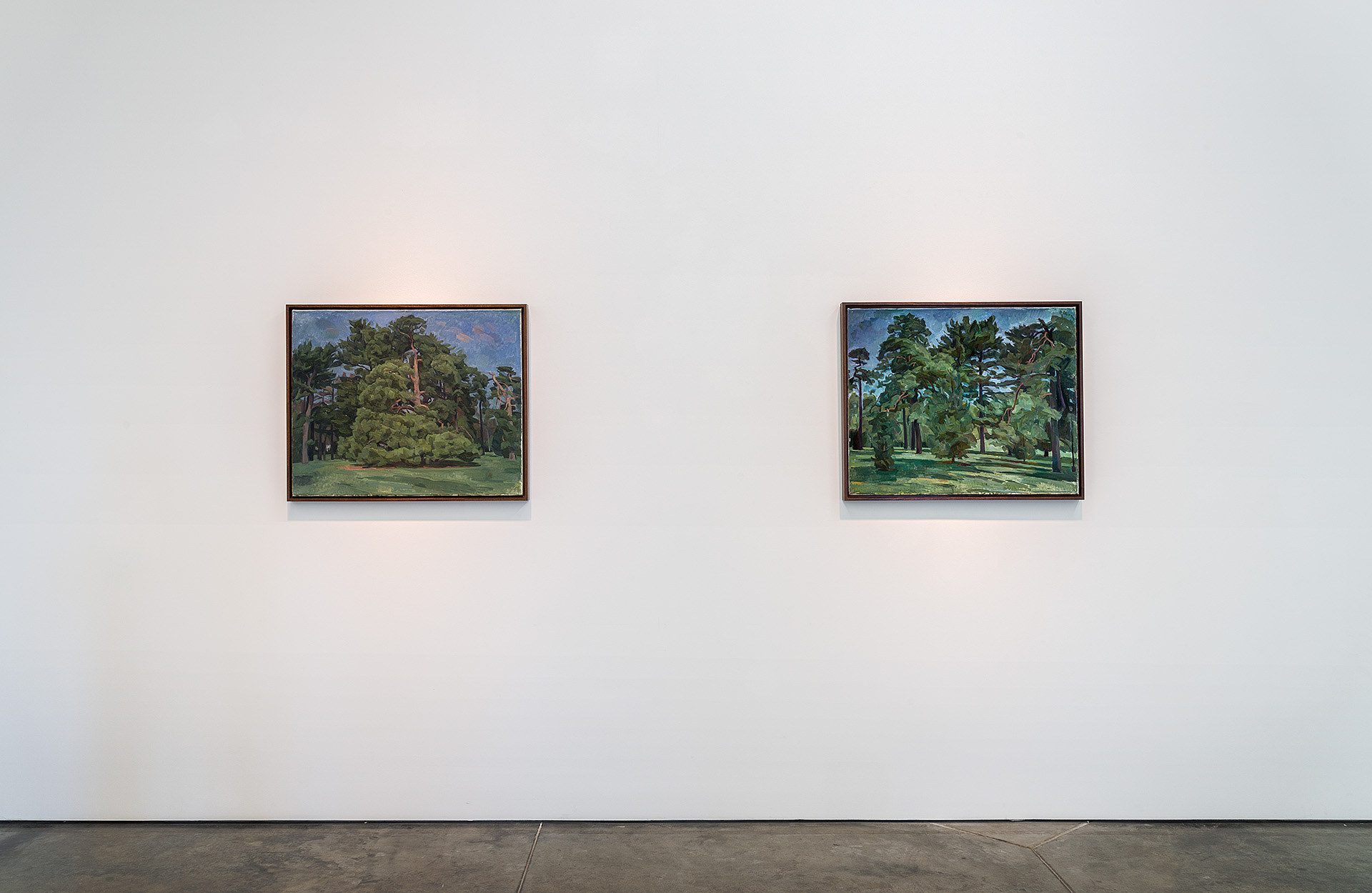 Two framed landscape paintings depicting dense, verdant forests with tall trees and green foliage hang on a white gallery wall. Both works feature naturalistic rendering in muted, earthy tones with blue skies visible above the tree canopies, positioned symmetrically across the expansive wall space.