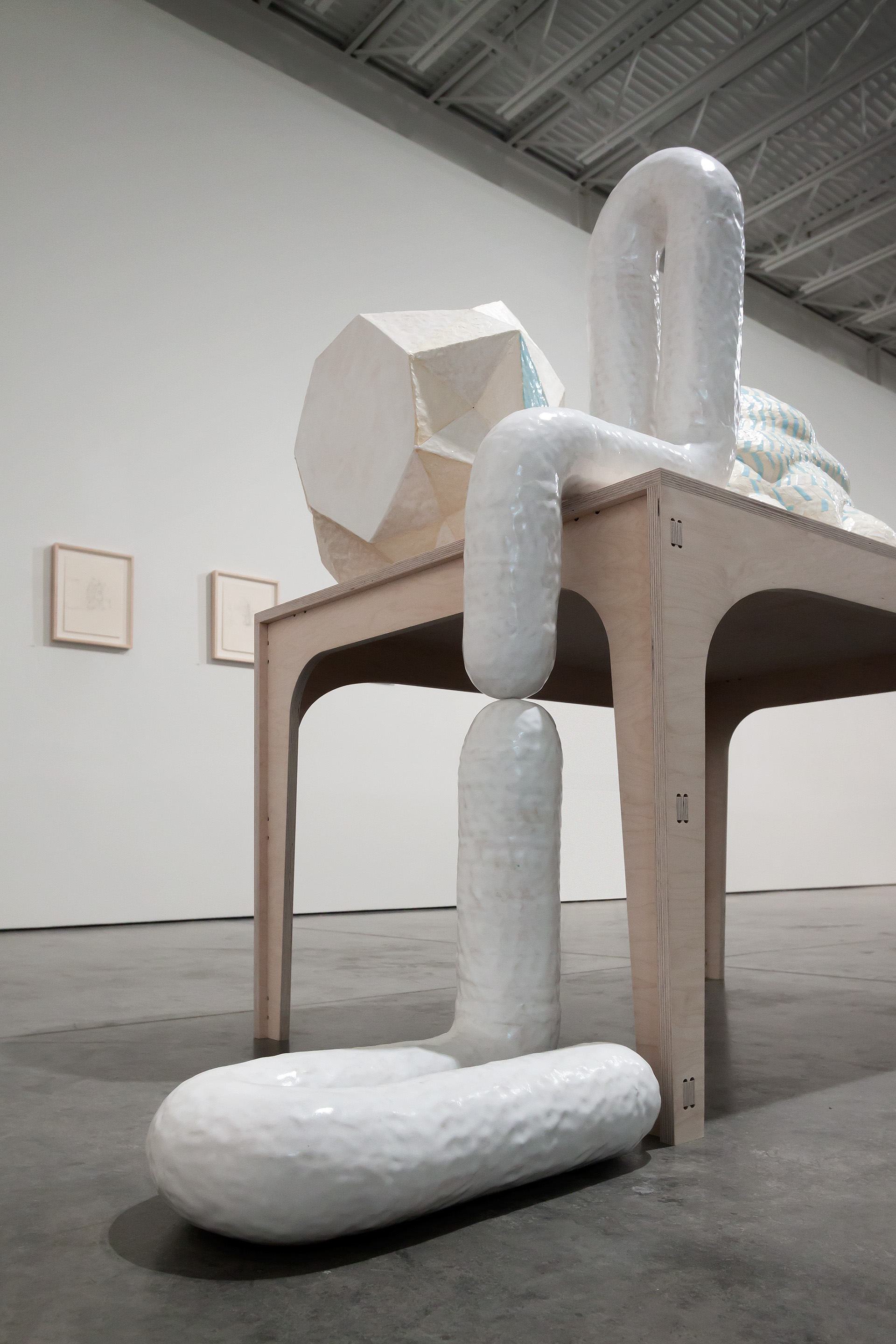 A large-scale sculptural installation featuring white glazed ceramic forms stacked vertically within and around a wooden frame structure. Organic, tubular shapes in varying sizes are balanced on and extending from a wooden platform, with an angular geometric form mounted at the top, creating an abstract composition that explores the relationship between containment, form, and the human scale.