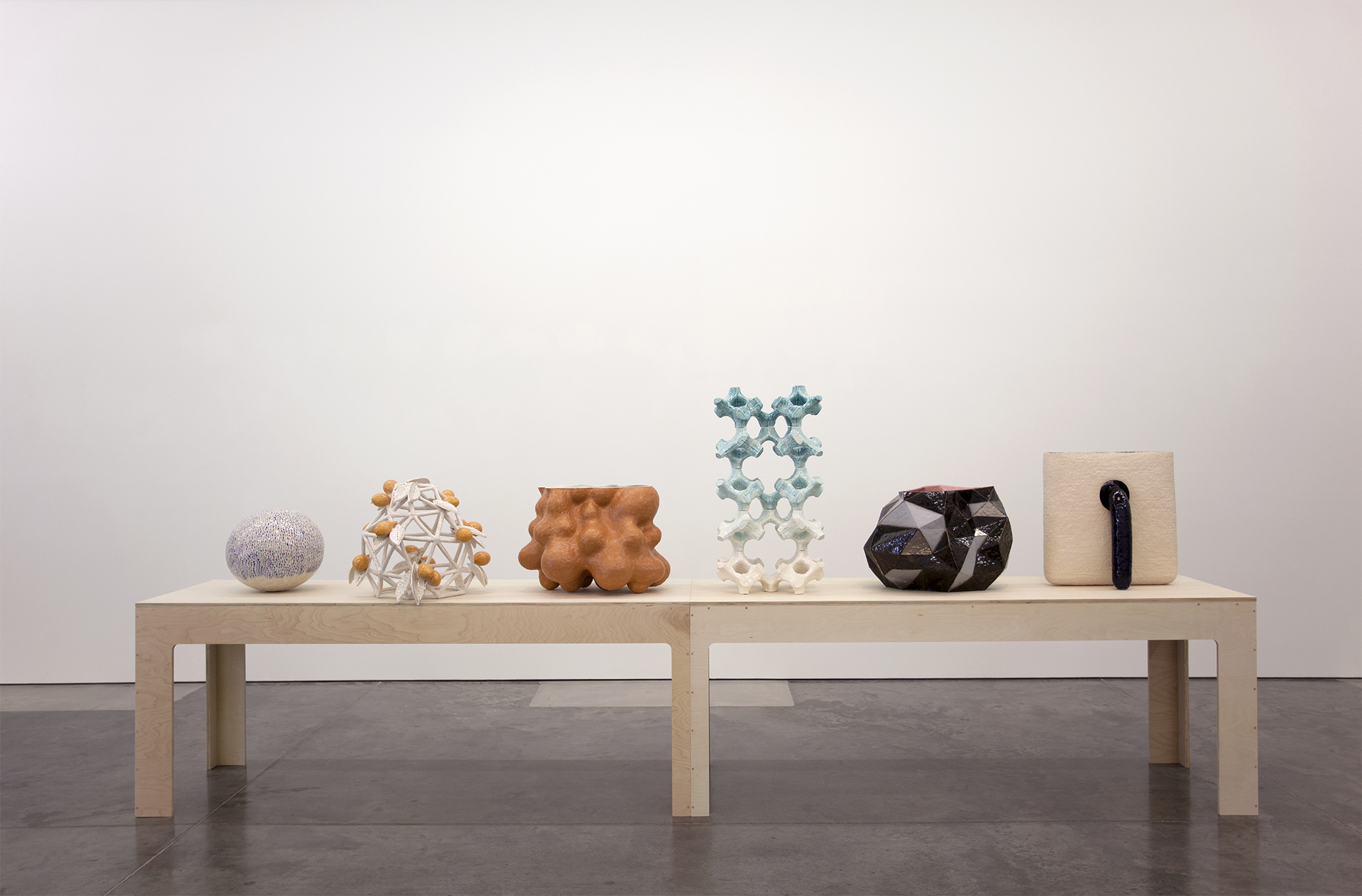 Six sculptural objects arranged in a horizontal still life on a light wood platform: a textured spherical form in lavender, a geometric gold and white faceted structure, an organic brown bulbous shape, a pale blue-green symmetrical form with void spaces, a dark faceted black and blue geometric object, and a cream-colored rectangular form with a black curved element. Clean gallery setting with white walls and gray floor.