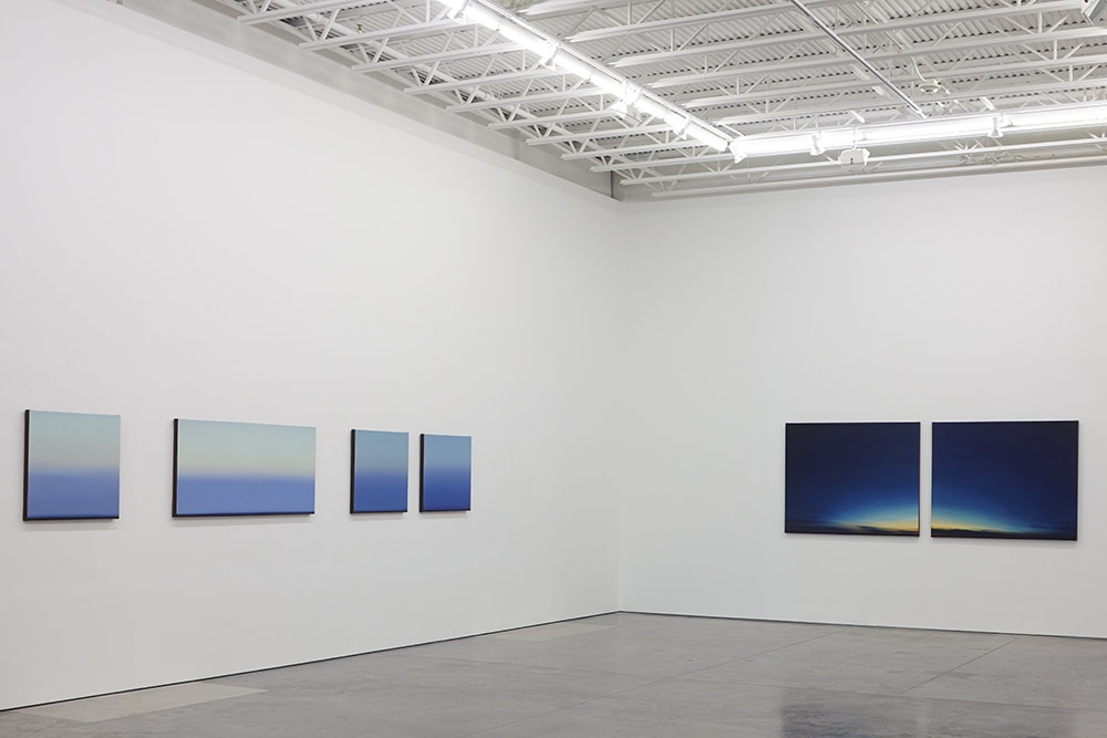 Five framed paintings hung on a white gallery wall depicting horizontal landscape compositions with gradient transitions from light upper sections to deeper blue tones, some with hints of golden light along the horizon; arranged in a sparse grouping with two diptychs on the right side featuring darker, more saturated color fields.