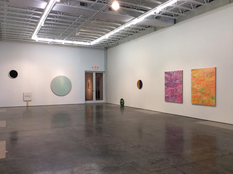 Contemporary gallery interior with polished concrete floor featuring circular mirror-like sculptures in muted green and black tones on white walls, alongside two vibrant abstract paintings in hot pink and golden yellow with densely patterned surfaces on the right wall.